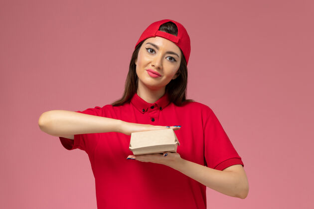 送货正面图：身穿红色制服斗篷的年轻女快递员 手上拿着一个小小的快递食品包 放在淡粉色的墙上手人微笑
