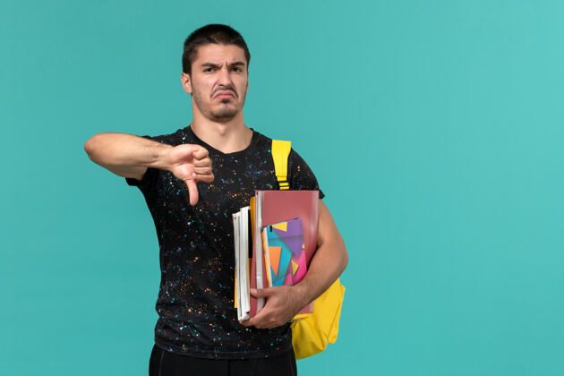 人身穿深色t恤 背着黄色背包 拿着文案和文件的男学生的正面图 蓝色墙上学院字帖书