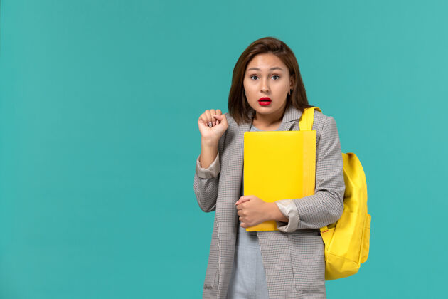 大学身穿灰色夹克 背着黄色背包 在浅蓝色墙上拿着文件的女学生的正视图人女学生学校