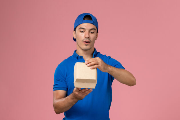 包裹正面图：身穿蓝色制服披肩的年轻男性快递员在粉红色的墙上拿着小包裹成人快递员正面