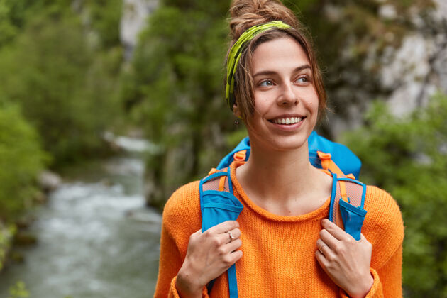 背包愁眉苦脸的快乐女人在雄伟的地方旅行 愉快地看着远方 穿着休闲的橙色毛衣 背着背包岩石休闲背包客
