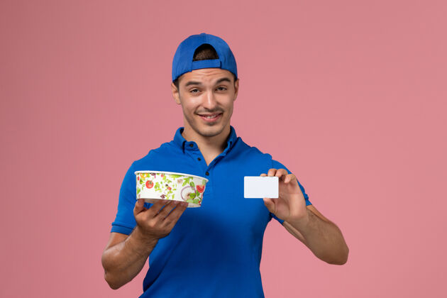 粉色正面图身穿蓝色制服披肩的年轻男性信使手持白色卡片和浅粉色墙上的圆形投递碗正面送货人