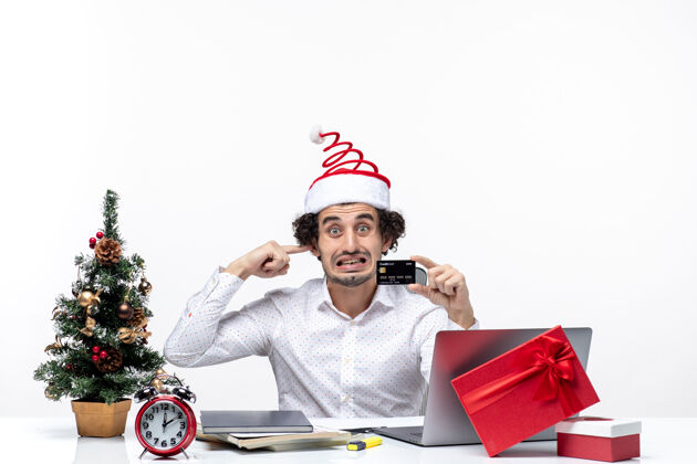 耳朵小胡子生气的商人戴着圣诞老人的帽子 手里拿着银行卡 在办公室里闭着耳朵办公室成年人胡子