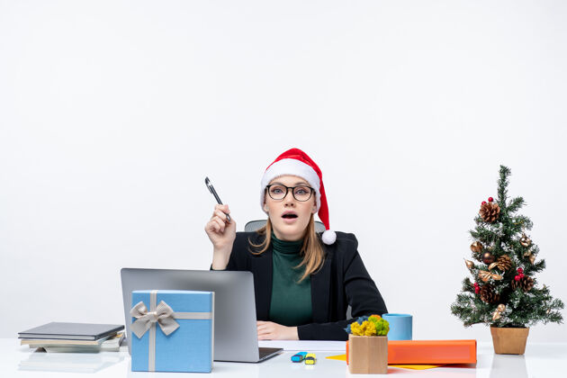 工作好奇的金发女人 戴着圣诞老人的帽子 坐在一张桌子旁 桌子上放着一棵圣诞树和一份白色背景的礼物圣诞老人女人好奇