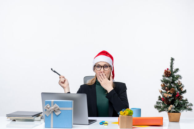 惊讶一个戴着圣诞老人帽子的金发女人坐在桌子旁 桌子上放着一棵圣诞树和一份白色背景的礼物圣诞树帽子笔记本电脑