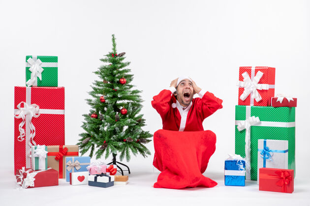 坐紧张的年轻人打扮成圣诞老人 拿着礼物和装饰好的圣诞树坐在白色背景的地上装饰礼物礼物