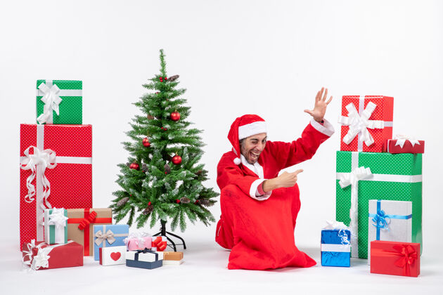 礼物微笑有趣的年轻人打扮成圣诞老人与礼物和装饰圣诞树坐在地上指着东西在左边的白色背景左边年轻人地