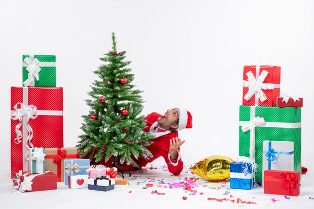 惊喜圣诞气氛与惊喜的圣诞老人躲在圣诞树后 在白色背景上不同颜色的礼物附近圣诞礼物季节