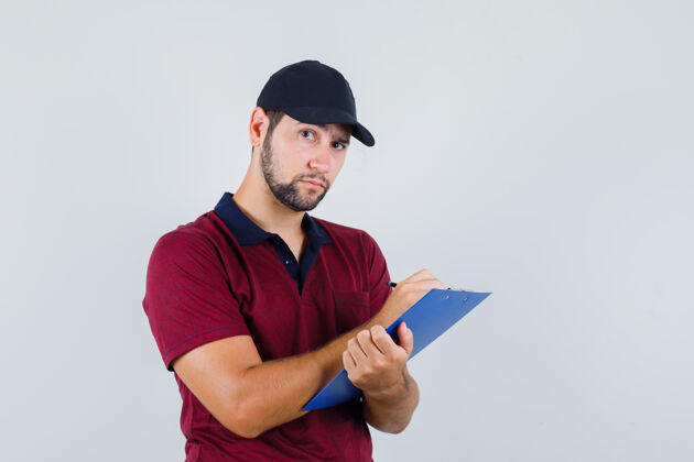 黑年轻的男性穿着红色t恤 戴着黑色帽子在笔记本上写着什么 看上去很专注 正对着前方笔记本休闲年轻