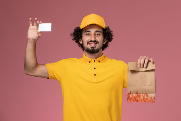 送货正面图身穿黄色制服的男快递员拿着食品包和塑料卡在粉红色的墙上持有黄色包裹