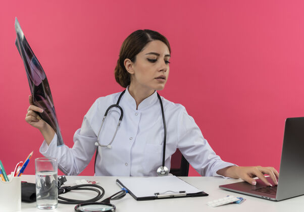 笔记本电脑想想年轻的女医生穿着医用长袍 带听诊器 坐在办公桌上 拿着医用工具 拿着x光片 用笔记本电脑 还有复印空间X光医生思考