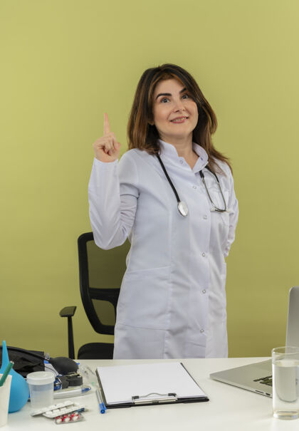 医生高兴的中年女医生穿着带听诊器的医用长袍 站在办公桌后面 拿着带医疗工具的笔记本电脑 指着隔离的绿色墙壁 还有复印空间高兴中年办公桌
