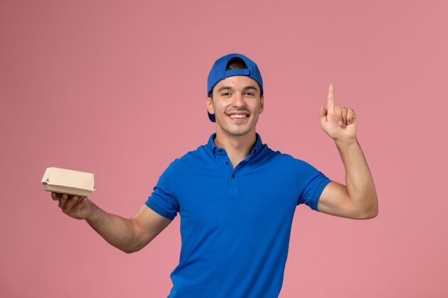 食物正面图：身穿蓝色制服披肩的年轻男性快递员手拿着小快递食品包站在粉红色的墙上粉色制服服务