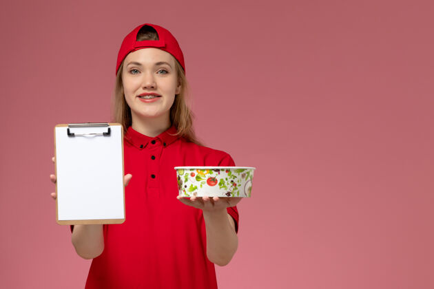 记事本正面图：身穿红色制服和披风的年轻女快递员拿着递碗和记事本 微笑着站在粉红色的墙上红色碗女人