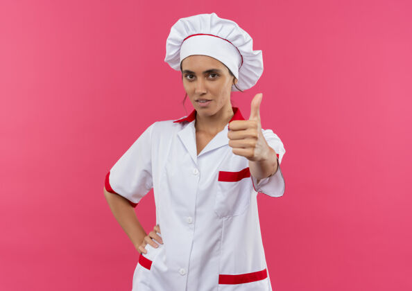 女年轻的女厨师穿着厨师制服 竖起大拇指 将手放在臀部 放在隔离的粉色墙壁上 留有复印空间穿厨师手