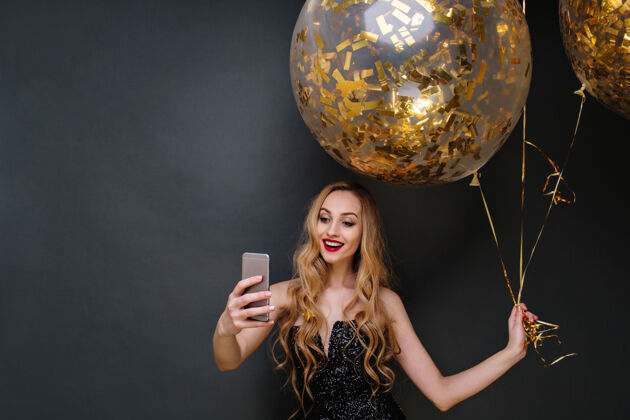 气球迷人的快乐的年轻迷人的女人穿着黑色的豪华礼服 长卷曲的金发自拍的大气球充满了金色的镀金庆祝现代党金色微笑电话