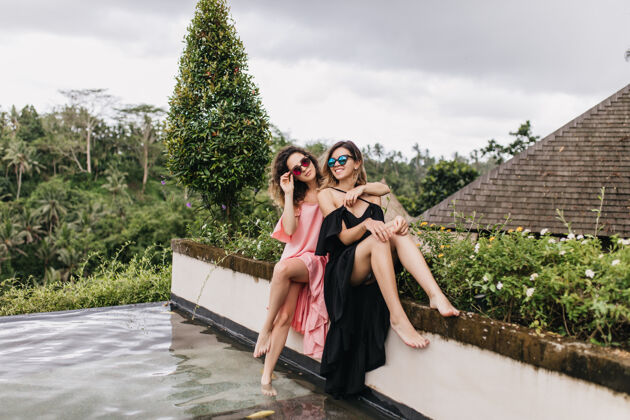 成人赤脚黑衣女子与妹妹在游泳池旁合影戴着墨镜的女模特在巴厘岛度假放松美丽花园奢华