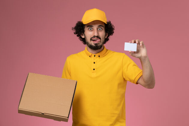 黄色正面图身穿黄色制服的男快递员拿着食品递送箱和卡片在粉色墙上生意男服务